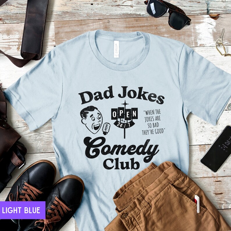 May include: Light blue t-shirt with a black graphic of a man with a microphone and the text "Dad Jokes Comedy Club" with a sign that says "Open 24/7" and the text "When the jokes are so bad they're good."