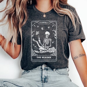 May include: A dark gray t-shirt with a white graphic of a tarot card featuring a skeleton reading a book. The card is titled "The Reader".