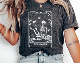 May include: A dark gray t-shirt with a white graphic of a tarot card featuring a skeleton reading a book. The card is titled "The Reader".