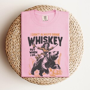 May include: Pink t-shirt with a graphic of a woman riding a bear and holding a bottle of whiskey. The text on the shirt reads "I don't always drink whiskey, but when I do..."