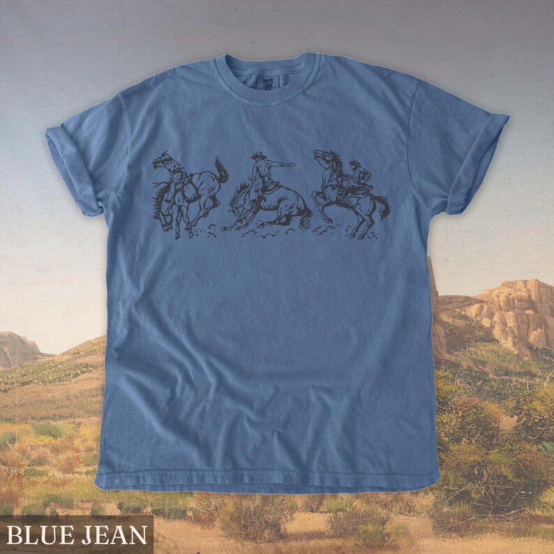 Puede incluir: Camiseta azul vaquero con un gr&aacute;fico negro de un vaquero montando un bronco. El gr&aacute;fico muestra tres escenas secuenciales del vaquero intentando mantenerse sobre el caballo. El texto "BLUE JEAN" est&aacute; en la parte inferior de la imagen.