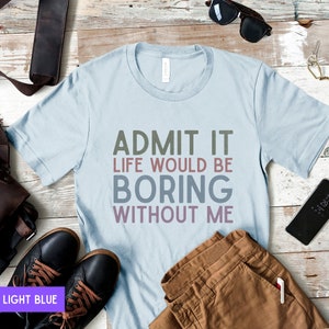 May include: Light blue t-shirt with the text "ADMIT IT LIFE WOULD BE BORING WITHOUT ME" in a multi-colored font. The shirt is displayed with brown pants, black shoes, and sunglasses on a wooden surface.