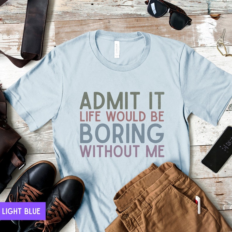 May include: Light blue t-shirt with the text "ADMIT IT LIFE WOULD BE BORING WITHOUT ME" in a multi-colored font. The shirt is displayed with brown pants, black shoes, and sunglasses on a wooden surface.