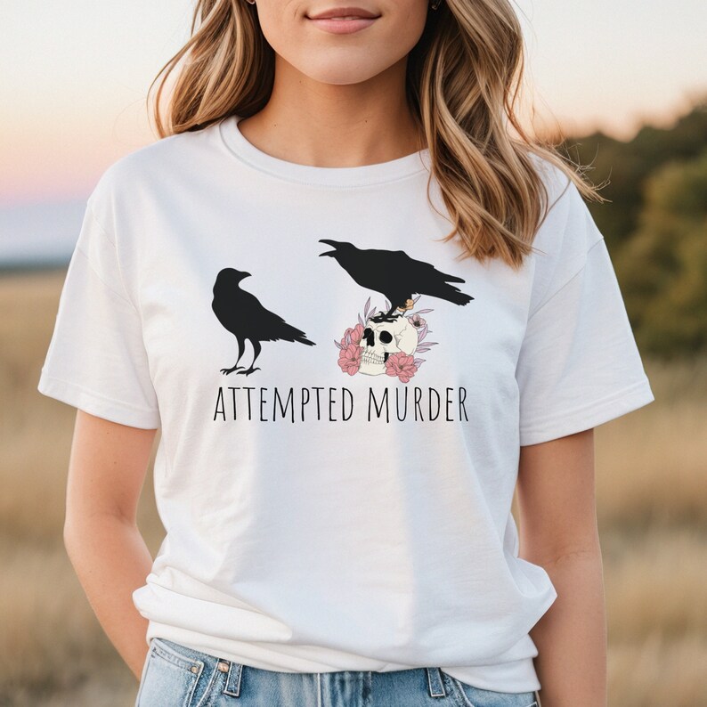 以下が含まれることがあります： 2 羽の黒いカラス、ピンクの花で飾られた骸骨、そして「ATTEMPTED MURDER」のテキストが特徴の白いTシャツ。 デザインは中央に配置され、ゴシックまたはハロウィーンのテーマを作成します。
