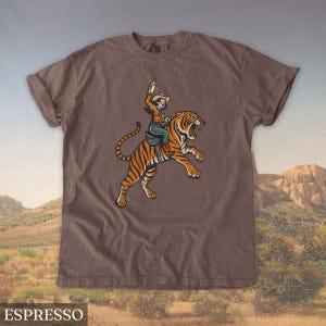 May include: Espresso-colored t-shirt featuring a cartoon illustration of a person riding a tiger. The person is wearing a cowboy hat and holding a bottle. The tiger is orange and black. The word "ESPRESSO" is at the bottom of the shirt.