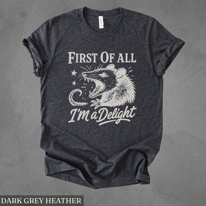 May include: Dark grey heather t-shirt with the text "FIRST OF ALL I'M a Delight" and a graphic of a possum with its mouth open. The possum is in black and white, with stars and a curved line design.