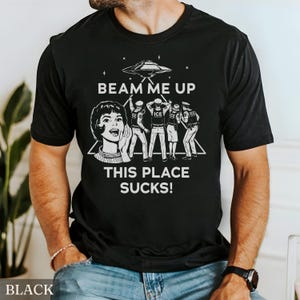 May include: Black t-shirt with a white graphic design. The design features a retro illustration of a woman shouting, a UFO, and figures with the text "BEAM ME UP THIS PLACE SUCKS!". The shirt is a crew neck style.