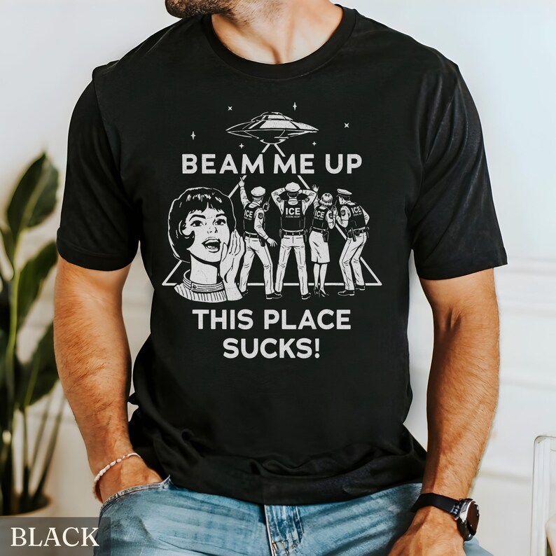 May include: Black t-shirt with a white graphic design. The design features a retro illustration of a woman shouting, a UFO, and figures with the text "BEAM ME UP THIS PLACE SUCKS!". The shirt is a crew neck style.