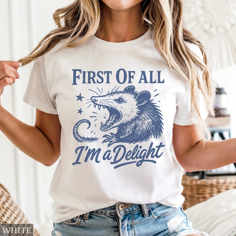 May include: White t-shirt with a blue graphic of an opossum with an open mouth and the text "FIRST OF ALL I'M a Delight". The design includes two stars and a curved tail.