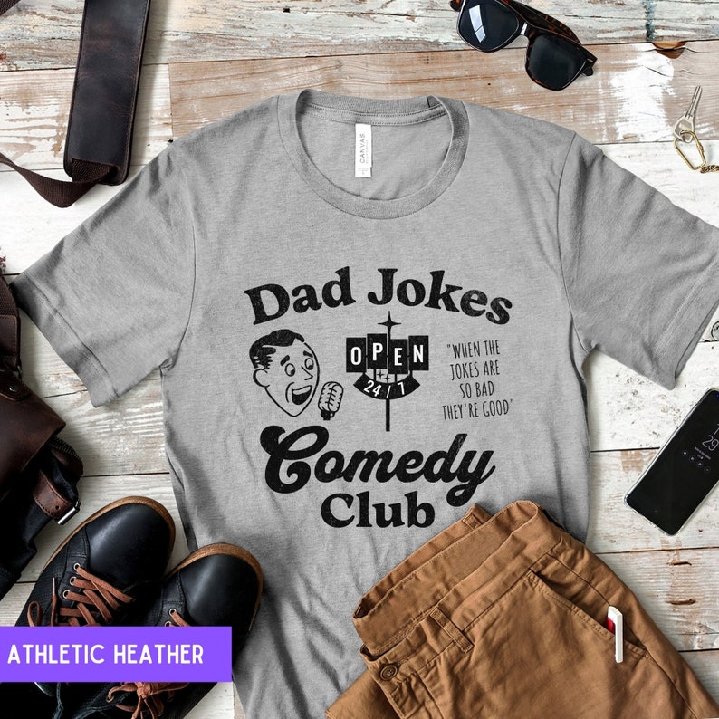 May include: A gray t-shirt with a black graphic design that says "Dad Jokes Comedy Club" with a cartoon man holding a microphone and a sign that says "Open 24/7". The text below the sign says "When the jokes are so bad they're good."