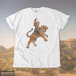 May include: White t-shirt featuring a vintage-style illustration of a person riding a tiger, holding a bottle. The tiger is orange and black, with the rider wearing a cowboy hat and holding a bottle with red liquid. The word "WHITE" is at the bottom.