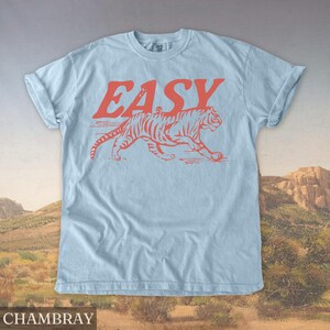 Op de afbeelding: Lichtblauw T-shirt met het woord "EASY" in rode letters boven een rode tijgerafbeelding. Het shirt is gemaakt van zacht materiaal en heeft opgerolde mouwen. Het woord "CHAMBRAY" staat onderaan de afbeelding.