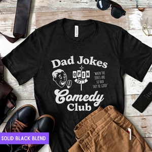 May include: Black t-shirt with a white graphic design that says "Dad Jokes Comedy Club" with a cartoon man holding a microphone and a sign that says "Open 24/7". The text below the sign says "When the jokes are so bad they're good."