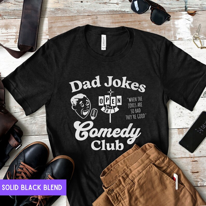 May include: Black t-shirt with a white graphic design that says "Dad Jokes Comedy Club" with a cartoon man holding a microphone and a sign that says "Open 24/7". The text below the sign says "When the jokes are so bad they're good."