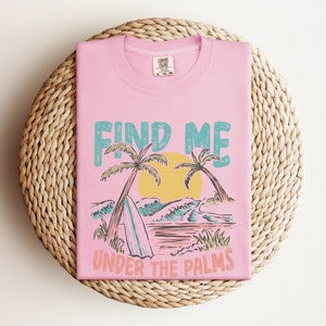 May include: Pink t-shirt with a graphic design of palm trees, waves, and a surfboard. The text "Find Me Under The Palms" is printed on the shirt.
