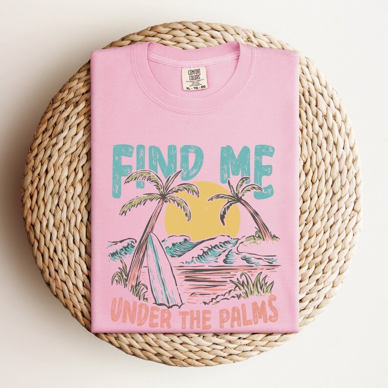 May include: Pink t-shirt with a graphic design of palm trees, waves, and a surfboard. The text "Find Me Under The Palms" is printed on the shirt.