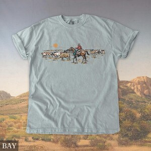 May include: A light blue t-shirt featuring a cowboy on horseback herding cattle under a setting sun. The graphic is in a vintage style, with a desert landscape background. The shirt has rolled-up sleeves and a relaxed fit.