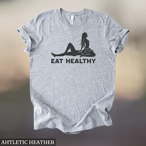 May include: Heather gray t-shirt with rolled sleeves. The shirt features a black graphic of a silhouette of a person lying down with another person sitting on top. The text "EAT HEALTHY" is below the graphic.