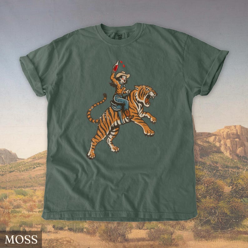 May include: Moss green t-shirt featuring a graphic of a person riding a tiger, holding a bottle. The person is wearing a cowboy hat and jeans. The tiger is orange with black stripes. The shirt has a relaxed fit.