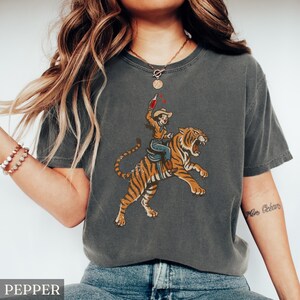 May include: Pepper-colored t-shirt featuring a graphic of a cowboy riding a tiger, holding a bottle. The tiger is orange and black, with the cowboy wearing a hat, blue jeans, and a brown shirt. The shirt has a vintage, worn look.