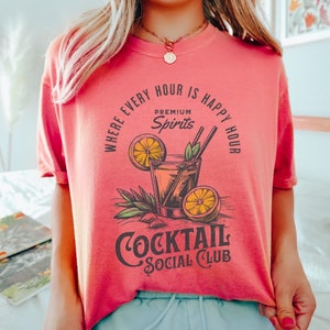 May include: A pink t-shirt with a graphic of a cocktail glass with a lemon wedge and the text "Where every hour is happy hour, Premium Spirits, Cocktail Social Club".