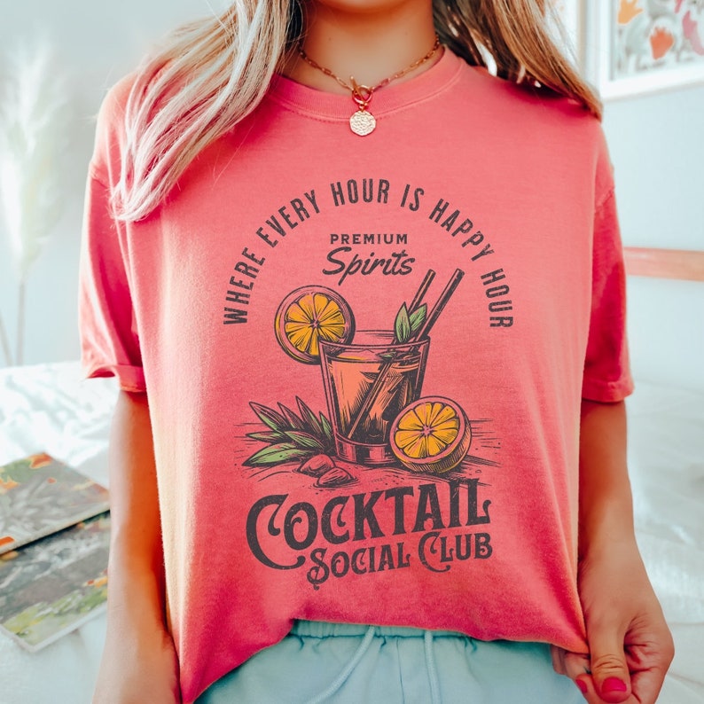 May include: A pink t-shirt with a graphic of a cocktail glass with a lemon wedge and the text "Where every hour is happy hour, Premium Spirits, Cocktail Social Club".
