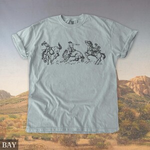 Puede incluir: Camiseta azul claro con una ilustraci&oacute;n en blanco y negro de vaqueros montando caballos salvajes. El gr&aacute;fico muestra tres escenas de rodeo en secuencia. La palabra "BAY" est&aacute; impresa en la parte inferior de la camiseta.