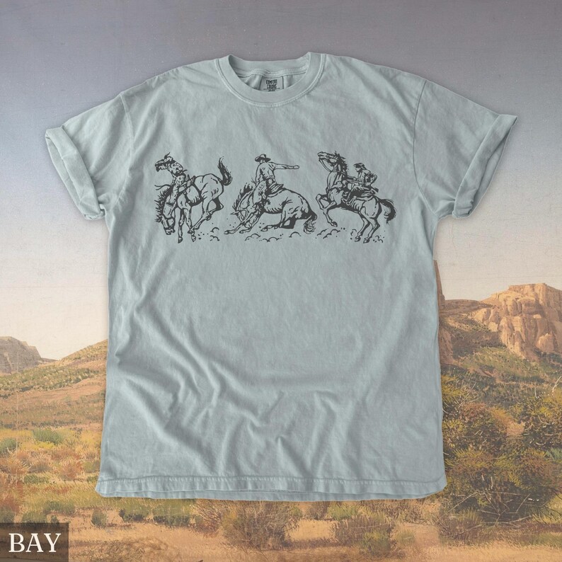 Puede incluir: Camiseta azul claro con una ilustraci&oacute;n en blanco y negro de vaqueros montando caballos salvajes. El gr&aacute;fico muestra tres escenas de rodeo en secuencia. La palabra "BAY" est&aacute; impresa en la parte inferior de la camiseta.