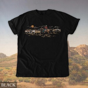 May include: Black t-shirt featuring a cowboy on horseback herding cattle under a setting sun. The graphic is in shades of brown, tan, and orange, depicting a classic Western scene. The word "BLACK" is printed at the bottom of the shirt.