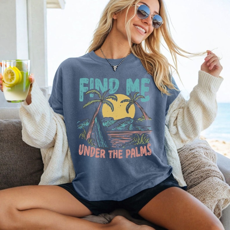 May include: A blue t-shirt with a graphic of palm trees, a sunset, and waves. The text "Find Me Under The Palms" is printed on the shirt.