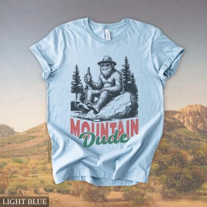 May include: Light blue t-shirt featuring a cartoon Bigfoot figure wearing a hat and holding a bottle, with the text "MOUNTAIN Dude" in red and green. The design includes pine trees and a rock. The shirt is made of a soft material.