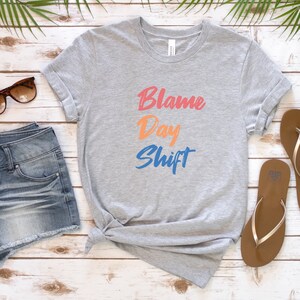 May include: A gray t-shirt with the text "Blame Day Shift" in colorful lettering. The shirt is folded and laid on a wooden surface with a pair of denim shorts and sandals.