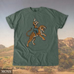 May include: Moss green t-shirt featuring a graphic of a person riding a tiger, holding a bottle. The person is wearing a cowboy hat, shirt, and jeans. The tiger has orange and black stripes. The word "MOSS" is in a white box at the bottom.
