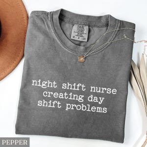 May include: A gray Comfort Colors t-shirt with the text "night shift nurse creating day shift problems" in white. A silver necklace with a small pendant is on the shirt. A brown hat is partially visible in the upper left corner.