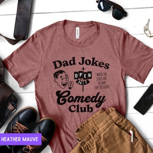 May include: Heather mauve t-shirt with a black graphic design that says "Dad Jokes Comedy Club" with a cartoon man holding a microphone and a sign that says "Open 24/1". The text below the sign says "When the jokes are so bad they're good."