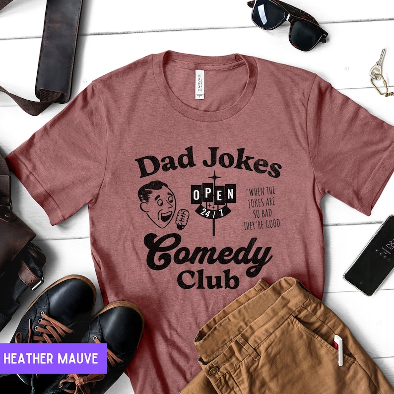May include: Heather mauve t-shirt with a black graphic design that says "Dad Jokes Comedy Club" with a cartoon man holding a microphone and a sign that says "Open 24/1". The text below the sign says "When the jokes are so bad they're good."