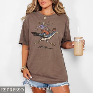 May include: Espresso-colored t-shirt featuring a cartoon illustration of a cowboy riding a roadrunner. The cowboy wears a purple shirt and hat, holding a lasso. The roadrunner is blue, black, and white. The word "ESPRESSO" is at the bottom.