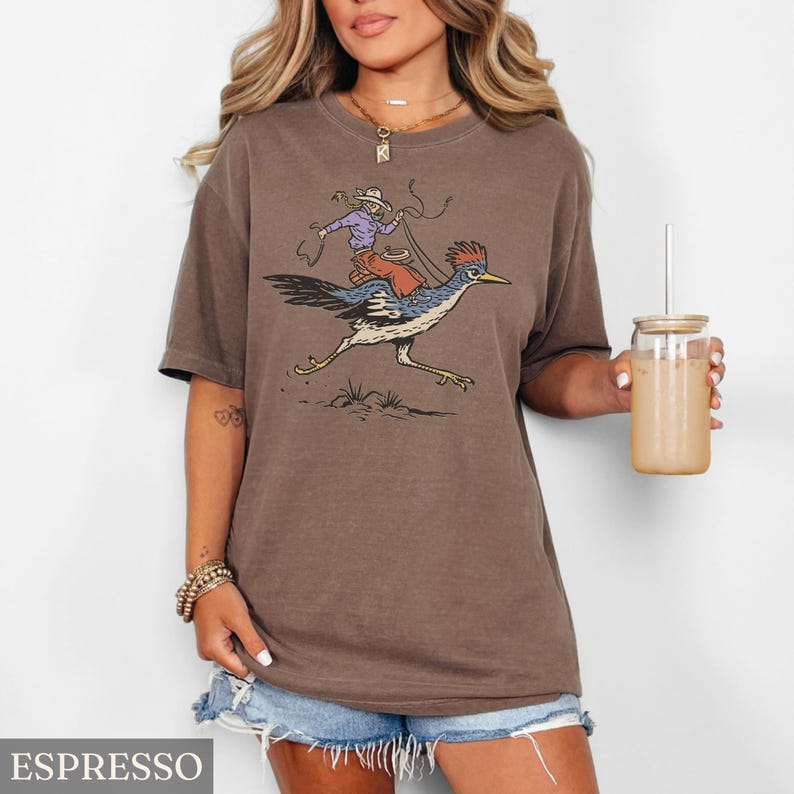 May include: Espresso-colored t-shirt featuring a cartoon illustration of a cowboy riding a roadrunner. The cowboy wears a purple shirt and hat, holding a lasso. The roadrunner is blue, black, and white. The word "ESPRESSO" is at the bottom.