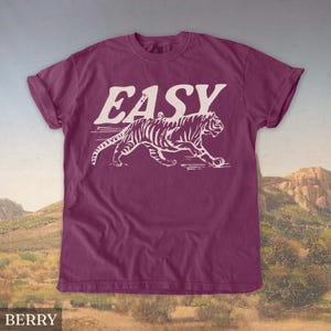 Op de afbeelding: Bessenkleurig T-shirt met het woord "EASY" in grote, witte blokletters boven een witte tijgerillustratie. De tijger loopt, met zijn strepen zichtbaar. Het shirt heeft korte mouwen en een ronde hals.