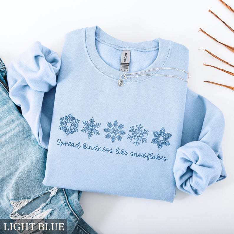 May include: A light blue sweatshirt with a crew neck and long sleeves. The design features five blue snowflakes and the text "Spread kindness like snowflakes." A silver necklace with a pendant is draped across the sweatshirt.