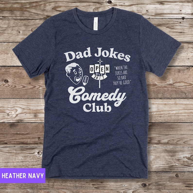 May include: A navy blue t-shirt with a white graphic of a cartoon man holding a microphone in front of a sign that says "Dad Jokes Comedy Club" and "When the jokes are so bad they're good".