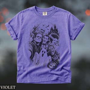 May include: A violet short-sleeve t-shirt featuring a dark graphic print of classic horror movie monsters including Frankenstein, Dracula, a mummy, a ghost, and a werewolf in front of a castle. The inner neck label displays the text Comfort Colors.
