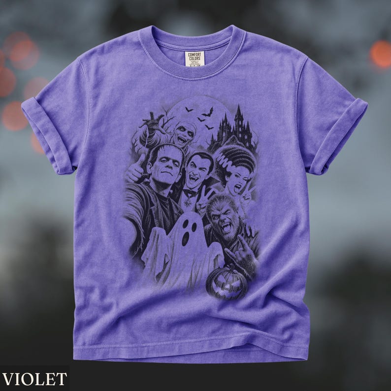 May include: A violet short-sleeve t-shirt featuring a dark graphic print of classic horror movie monsters including Frankenstein, Dracula, a mummy, a ghost, and a werewolf in front of a castle. The inner neck label displays the text Comfort Colors.