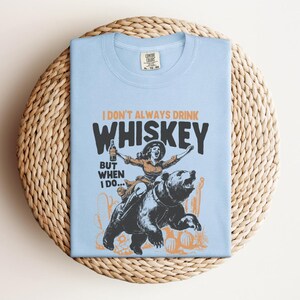 May include: Light blue t-shirt with a graphic of a woman riding a bear and holding a bottle of whiskey. The text on the shirt reads "I don't always drink whiskey, but when I do..."