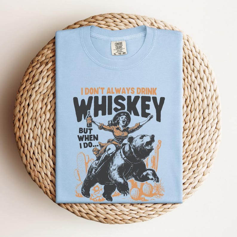 May include: Light blue t-shirt with a graphic of a woman riding a bear and holding a bottle of whiskey. The text on the shirt reads "I don't always drink whiskey, but when I do..."