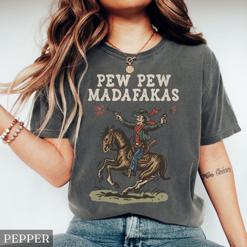 Puede incluir: Camiseta gris oscuro con el texto "PEW PEW MADAFAKAS" sobre un dibujo de un vaquero a caballo disparando con pistolas. El vaquero lleva un pa&ntilde;uelo rojo y un sombrero. La camiseta es de corte holgado.