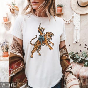 May include: White t-shirt featuring a vintage-style illustration of a cowgirl riding a tiger, holding a bottle. The tiger is orange with black stripes. The cowgirl is wearing a hat, blue jeans, and a brown jacket. The word "WHITE" is at the bottom.