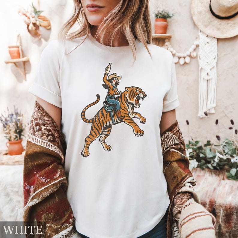May include: White t-shirt featuring a vintage-style illustration of a cowgirl riding a tiger, holding a bottle. The tiger is orange with black stripes. The cowgirl is wearing a hat, blue jeans, and a brown jacket. The word "WHITE" is at the bottom.