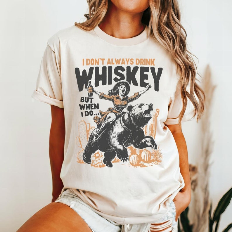 May include: A beige t-shirt with a graphic print of a woman riding a bear. The text on the shirt reads "I don't always drink whiskey, but when I do..."