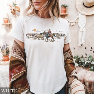 May include: White t-shirt featuring a Western-themed graphic. The design depicts a cowboy on horseback herding cattle under a setting sun. The word "WHITE" is printed at the bottom of the shirt.